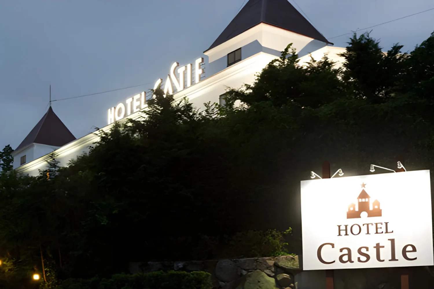 夕暮れの空を背に、屋根の「HOTEL CASTLE」の文字が灯るホテルキャッスルの外観。手前に城のロゴが入った白い案内看板。